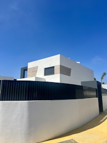 A modern, minimalist house with clean lines and geometric shapes. The exterior features a combination of white walls and light wooden panels. A high black fence surrounds the property, and there is a clear blue sky in the background.