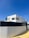 A modern, minimalist house with clean lines and geometric shapes. The exterior features a combination of white walls and light wooden panels. A high black fence surrounds the property, and there is a clear blue sky in the background.
