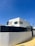 A modern, minimalist house with clean lines and geometric shapes. The exterior features a combination of white walls and light wooden panels. A high black fence surrounds the property, and there is a clear blue sky in the background.
