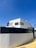 A modern, minimalist house with clean lines and geometric shapes. The exterior features a combination of white walls and light wooden panels. A high black fence surrounds the property, and there is a clear blue sky in the background.
