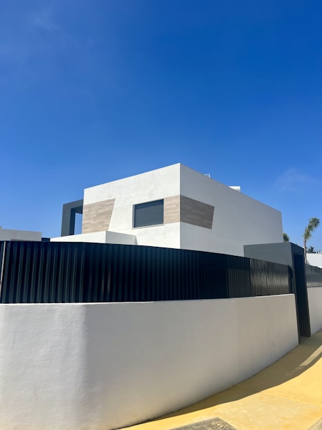 A modern, minimalist house with clean lines and geometric shapes. The exterior features a combination of white walls and light wooden panels. A high black fence surrounds the property, and there is a clear blue sky in the background.