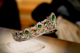 A luxurious tiara adorned with large, vibrant green gemstones and intricate patterns of sparkling diamonds. The tiara is elegantly displayed against a blurred background, enhancing its opulence and intricate craftsmanship.