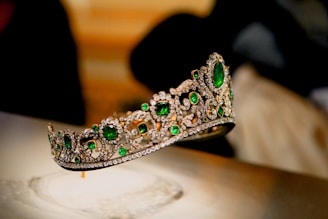A luxurious tiara adorned with large, vibrant green gemstones and intricate patterns of sparkling diamonds. The tiara is elegantly displayed against a blurred background, enhancing its opulence and intricate craftsmanship.