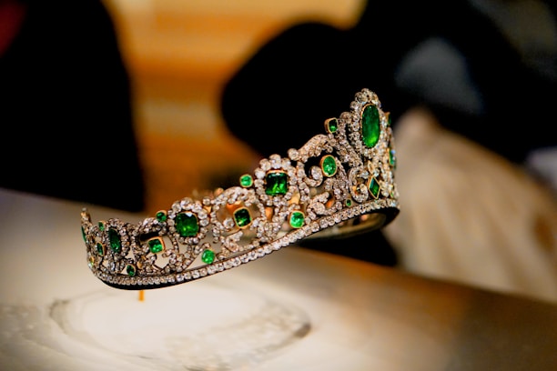 A luxurious tiara adorned with large, vibrant green gemstones and intricate patterns of sparkling diamonds. The tiara is elegantly displayed against a blurred background, enhancing its opulence and intricate craftsmanship.