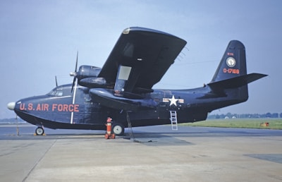 A dark-colored military aircraft marked with 'U.S. Air Force' in bold letters on its fuselage, stationed on an airfield. An aerial ladder is propped against the side of the aircraft near the wing. A red and white fire hydrant can be seen in front of the aircraft's nose. The background shows a grassy landscape under a clear blue sky.