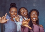 Three women are smiling and reaching towards the camera, creating a lively and engaging atmosphere. They appear happy and are dressed in colorful clothing against a plain background.