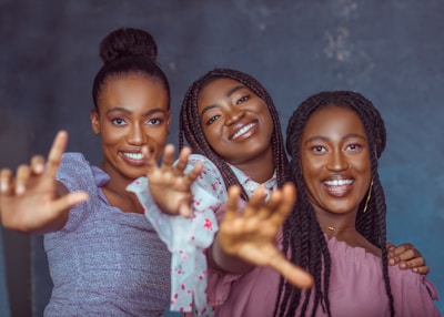 Three women are smiling and reaching towards the camera, creating a lively and engaging atmosphere. They appear happy and are dressed in colorful clothing against a plain background.