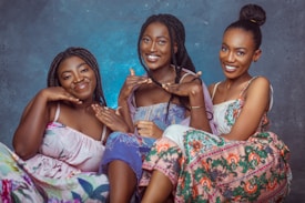 Three women are sitting closely together, wearing colorful dresses and smiling warmly. The background is a textured blue, adding a serene atmosphere to the composition. Their expressions and gestures convey a sense of joy and connection.