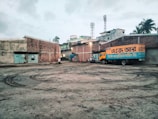 A truck with Bengali text painted on its side is parked in a large, open courtyard. The area is surrounded by brick buildings with some structures visible in the background, like two mobile towers. There are a few people and motorcycles gathered near the truck. The ground has tire marks and appears to be a little muddy.