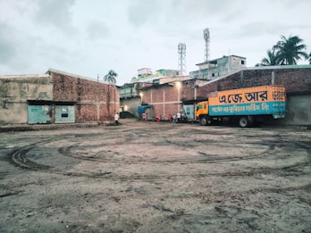 A truck with Bengali text painted on its side is parked in a large, open courtyard. The area is surrounded by brick buildings with some structures visible in the background, like two mobile towers. There are a few people and motorcycles gathered near the truck. The ground has tire marks and appears to be a little muddy.