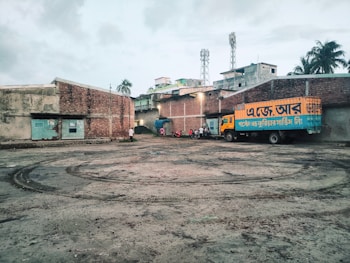 A truck with Bengali text painted on its side is parked in a large, open courtyard. The area is surrounded by brick buildings with some structures visible in the background, like two mobile towers. There are a few people and motorcycles gathered near the truck. The ground has tire marks and appears to be a little muddy.