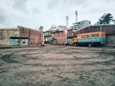 A truck with Bengali text painted on its side is parked in a large, open courtyard. The area is surrounded by brick buildings with some structures visible in the background, like two mobile towers. There are a few people and motorcycles gathered near the truck. The ground has tire marks and appears to be a little muddy.