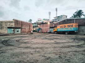A truck with Bengali text painted on its side is parked in a large, open courtyard. The area is surrounded by brick buildings with some structures visible in the background, like two mobile towers. There are a few people and motorcycles gathered near the truck. The ground has tire marks and appears to be a little muddy.