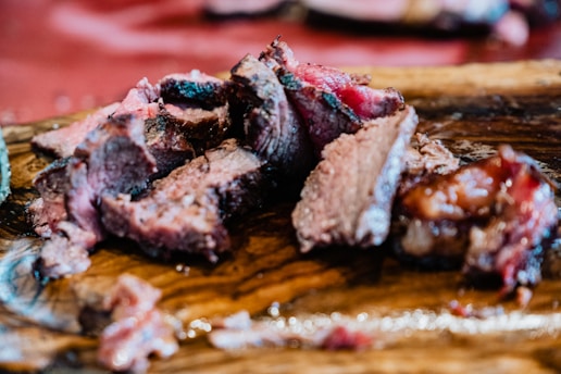 Juicy slices of cooked meat arranged on a wooden cutting board, showcasing a pinkish interior and charred edges. The surface below the meat appears to be glossy due to natural juices seeping from the cuts.