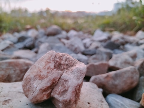 A close-up of a collection of stones with one prominently positioned in the foreground, displaying a rough texture and brownish color. The background is blurred with hints of greenery and an indistinct horizon, suggesting an outdoor setting during twilight.