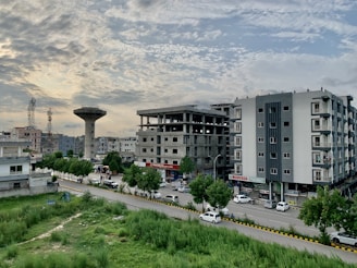A suburban area with residential buildings, including a multi-story construction in progress and a grey apartment complex. There are green trees lining the road, which is populated with parked cars and minimal traffic. The sky is partly cloudy, exhibiting a soft blend of light blue and gray tones.