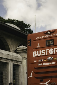 A red hop-on hop-off tour bus is partially visible with the back section in the foreground. The bus displays various flags and an advertisement for a sightseeing service in Istanbul. In the background, an old stone building with arched windows and lush green trees can be seen under a cloudy sky.