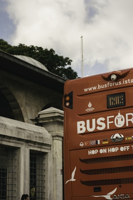A red hop-on hop-off tour bus is partially visible with the back section in the foreground. The bus displays various flags and an advertisement for a sightseeing service in Istanbul. In the background, an old stone building with arched windows and lush green trees can be seen under a cloudy sky.