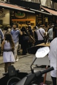 A bustling street market scene with multiple people walking and engaging in various activities. Shops line the street with visible signage, and a display of goods is evident in the foreground. The crowd includes individuals dressed in a range of attire, indicating a diverse environment.