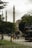A bustling street scene in a city with people walking near a historic mosque featuring two tall minarets. A red double-decker bus with tourists passes by, and lush green trees frame the scene.