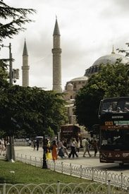 A bustling street scene in a city with people walking near a historic mosque featuring two tall minarets. A red double-decker bus with tourists passes by, and lush green trees frame the scene.