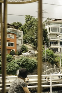A scenic view of a hillside residential area featuring several modern houses with balconies and large windows. The buildings are surrounded by lush, green foliage. Two cars are parked on a sloped driveway leading uphill. In the foreground, a person wearing a hat is visible, with their back turned, leaning on a railing. Near the water, a few other people are engaged in activities, possibly fishing.