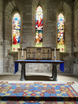 An ornate altar is adorned with a vibrant tapestry featuring religious motifs, situated beneath three intricate stained glass windows depicting biblical scenes. The stone walls add a rustic ambiance, complementing the sanctuary setting.