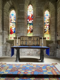 An ornate altar is adorned with a vibrant tapestry featuring religious motifs, situated beneath three intricate stained glass windows depicting biblical scenes. The stone walls add a rustic ambiance, complementing the sanctuary setting.
