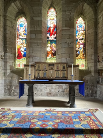 An ornate altar is adorned with a vibrant tapestry featuring religious motifs, situated beneath three intricate stained glass windows depicting biblical scenes. The stone walls add a rustic ambiance, complementing the sanctuary setting.