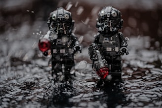 Two toy figures resembling pilots or soldiers in black armor are standing amidst a wet surface with water droplets around them. Both figures are holding objects with glowing red elements, possibly weapons or devices. The lighting and droplets suggest a dramatic, intense setting.