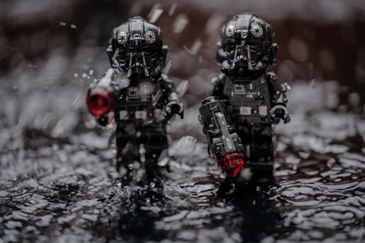 Two toy figures resembling pilots or soldiers in black armor are standing amidst a wet surface with water droplets around them. Both figures are holding objects with glowing red elements, possibly weapons or devices. The lighting and droplets suggest a dramatic, intense setting.