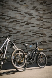 Two bicycles are parked on a pavement next to a brick wall. The bicycles have thick tires and are equipped with features typical for mountain or electric bikes. The wall is composed of rectangular bricks in varying shades of brown and beige. The setting is outdoors, with sunlight casting shadows.