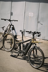 Two bicycles parked on a paved surface in front of a gray metal door. The bicycles, one black and one white, are modern and equipped with wide tires, suitable for off-road or mountain biking. The image captures a shadow on the ground, suggesting strong sunlight.
