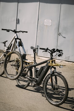 Two bicycles parked on a paved surface in front of a gray metal door. The bicycles, one black and one white, are modern and equipped with wide tires, suitable for off-road or mountain biking. The image captures a shadow on the ground, suggesting strong sunlight.