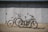 Two bikes are parked on a flat surface in front of a large, closed metal garage door. The bikes are of different styles and colors, one being a sleek black electric bike with a battery pack, and the other a white mountain bike with prominent front suspension.