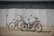 Two bikes are parked on a flat surface in front of a large, closed metal garage door. The bikes are of different styles and colors, one being a sleek black electric bike with a battery pack, and the other a white mountain bike with prominent front suspension.