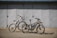 Two bikes are parked on a flat surface in front of a large, closed metal garage door. The bikes are of different styles and colors, one being a sleek black electric bike with a battery pack, and the other a white mountain bike with prominent front suspension.