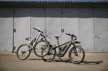 Two bikes are parked on a flat surface in front of a large, closed metal garage door. The bikes are of different styles and colors, one being a sleek black electric bike with a battery pack, and the other a white mountain bike with prominent front suspension.