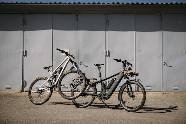 Two bikes are parked on a flat surface in front of a large, closed metal garage door. The bikes are of different styles and colors, one being a sleek black electric bike with a battery pack, and the other a white mountain bike with prominent front suspension.