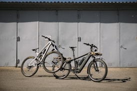 Two bikes are parked on a flat surface in front of a large, closed metal garage door. The bikes are of different styles and colors, one being a sleek black electric bike with a battery pack, and the other a white mountain bike with prominent front suspension.