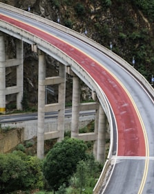 A curving bridge with a red surface and yellow lines supported by concrete pillars. It appears to be part of an elevated roadway, set against a backdrop of rocky terrain and greenery. Below, another road runs parallel with visible retaining walls.