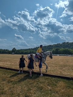 A person wearing a yellow shirt and helmet is mounted on a white horse on a vast grassy field under a partly cloudy sky. Three individuals, likely women in dark dresses, are gathered around the horse. In the background, there are tents and trees at the edge of the field, along with distant hills.