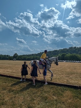 A person wearing a yellow shirt and helmet is mounted on a white horse on a vast grassy field under a partly cloudy sky. Three individuals, likely women in dark dresses, are gathered around the horse. In the background, there are tents and trees at the edge of the field, along with distant hills.