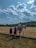 A person wearing a yellow shirt and helmet is mounted on a white horse on a vast grassy field under a partly cloudy sky. Three individuals, likely women in dark dresses, are gathered around the horse. In the background, there are tents and trees at the edge of the field, along with distant hills.