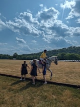 A person wearing a yellow shirt and helmet is mounted on a white horse on a vast grassy field under a partly cloudy sky. Three individuals, likely women in dark dresses, are gathered around the horse. In the background, there are tents and trees at the edge of the field, along with distant hills.