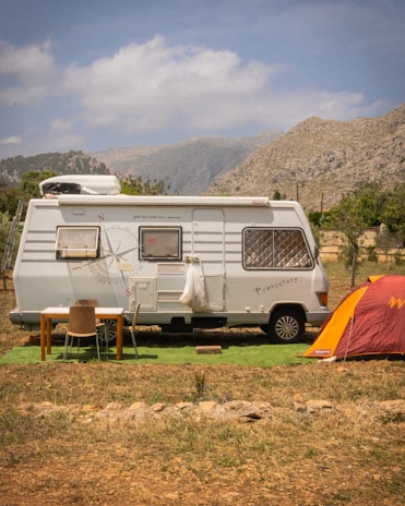 A white camper van is parked in a scenic, mountainous area with a cloudy sky. Next to the van, there is a small table and two chairs set up on a patch of artificial grass. To the right, an orange tent is pitched nearby. The background features rugged, rocky mountains and sparse greenery.