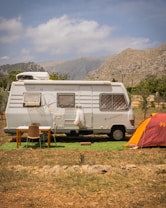 A white camper van is parked in a scenic, mountainous area with a cloudy sky. Next to the van, there is a small table and two chairs set up on a patch of artificial grass. To the right, an orange tent is pitched nearby. The background features rugged, rocky mountains and sparse greenery.