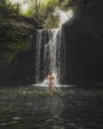 A person stands confidently on a bamboo raft with a wooden paddle on a serene body of water. A lush green forest surrounds a cascading waterfall in the background with sunlight filtering through the trees.