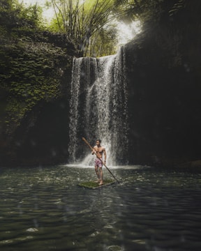 A person stands confidently on a bamboo raft with a wooden paddle on a serene body of water. A lush green forest surrounds a cascading waterfall in the background with sunlight filtering through the trees.