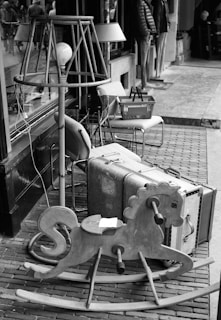 A collection of vintage items arranged outdoors along a sidewalk, including a large, old-fashioned suitcase, a wooden rocking horse, and a variety of lamps. Behind these items, a shop window and entrance are visible, with reflections of passersby on the glass. The scene has a nostalgic, antique feel.
